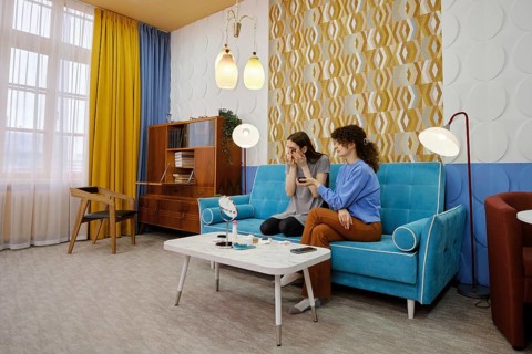 In a bright living room adorned with curtains, two friends sit on a blue sofa, chatting and