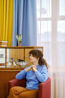 A woman with curly hair sits comfortably in a red chair, wearing a blue sweater and brown pants