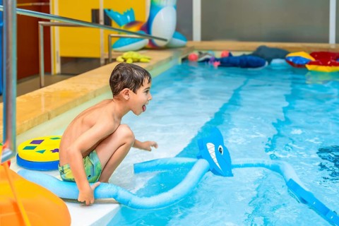 Happy child crouching at the edge of an indoor swimming pool, playing with a pool noodle shaped