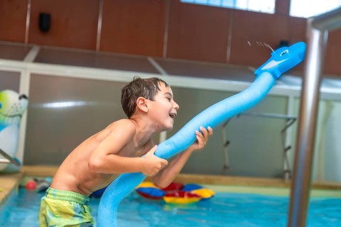 Happy child playing with a pool noodle shaped like a whale and spraying water in an indoor swimming