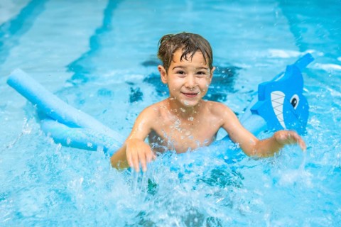 Smiling boy having fun in a swimming pool, playing with a pool noodle float shaped like a shark,