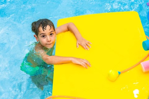 Young boy playing with a yellow floating mattress in a blue swimming pool during summer holidays