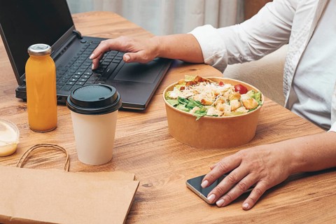 A woman is working on a laptop at home, there is food on the table, salad, coffee and juice,