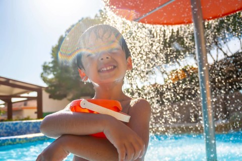 Smiling child holding a bucket and enjoying refreshing shower in a swimming pool on a sunny summer