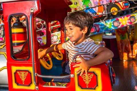 Young boy smiling while steering a vibrant red amusement park ride, surrounded by colorful
