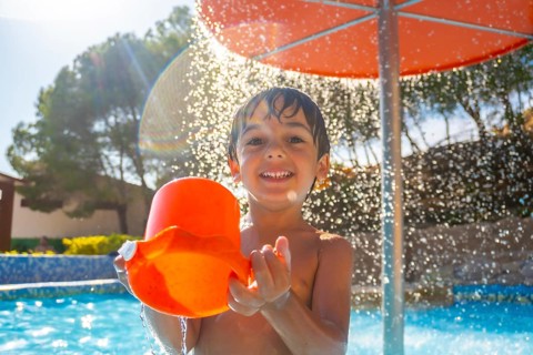 Smiling child enjoying a sunny day at the pool, holding an orange bucket under a refreshing water