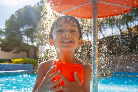 Smiling boy holding an orange water toy under a splash of water, enjoying a sunny day by the pool