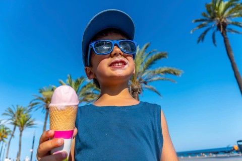 Young boy wearing sunglasses and a cap enjoys a strawberry ice cream cone on a sunny beach with