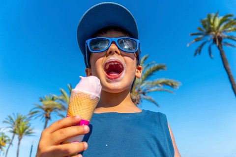 Young boy with sunglasses and cap enjoying a strawberry ice cream cone on a hot summer day with