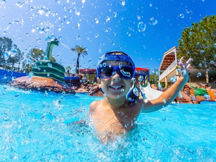 Happy boy wearing swimming goggles, splashing joyfully in a vibrant water park pool during summer