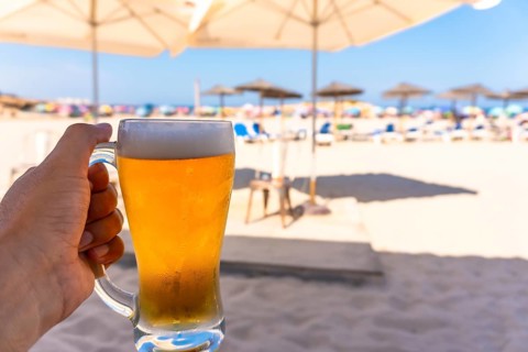 Tourist enjoying refreshing beer on sunny beach with blurred umbrellas and ocean, perfect summer