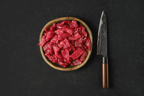 A wooden bowl holds freshly cut pieces of raw beef, showcasing a vibrant red color. Beside the