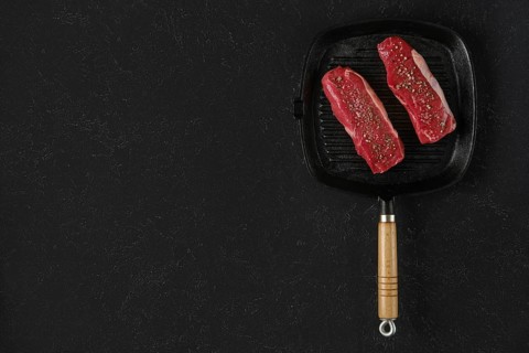 Two raw beef steaks on a preheated griddle pan, seasoned with spices, in a home kitchen setting.