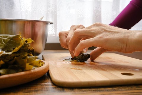 Hands delicately wrap seasoned filling in grape leaves on a wooden cutting board. Fresh ingredients