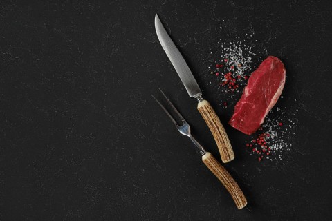 A knife and fork, with horn handles, rest beside a raw beef steak on a dark surface. Salt and pink