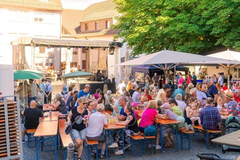 Visitors sit at beer tables at an open-air concert, surrounded by trees and parasols, 950 years