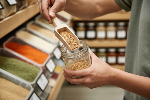 Hands filling small glass jar with grains using wooden scoop at bulk store. Concept of zero-waste