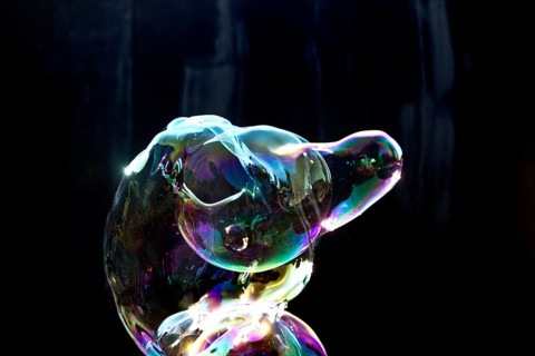 A shimmering and floating soap bubble in the colours of the rainbow against a black background,