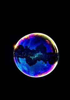 A shimmering and floating soap bubble in the colours of the rainbow against a black background,