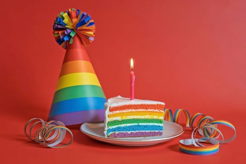 Rainbow birthday cake slice with rainbow party hat and streamers on bright red background.