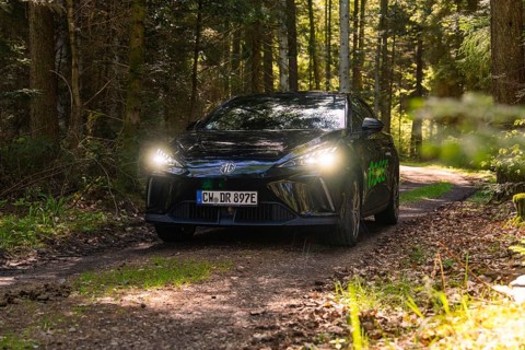 Black car with switched on headlights on a forest path, surrounded by trees, MG 4, Deer E- Car