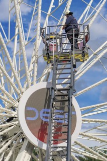 A worker stands on a ladder in front of a large Ferris wheel under a blue sky, fire brigade