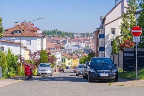 Urban street with cars, a no cycling sign, a scooter and a backdrop of houses under a clear sky,