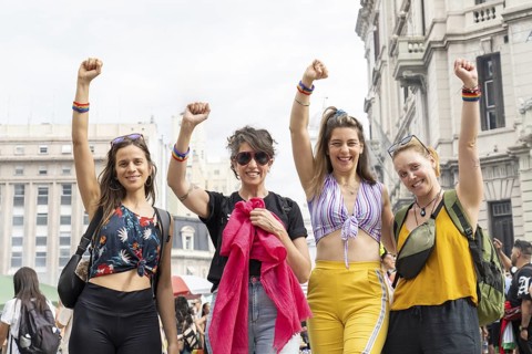 Four women are raising their arms, wearing rainbow bracelets and smiling, celebrating lgbtq rights