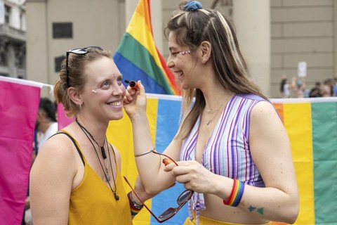 Two women are smiling and putting rainbow makeup on their faces at a gay pride parade, celebrating