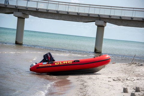 A DLRG lifeboat lies on the beach in front of a bridge with walkers and the sea in the background,