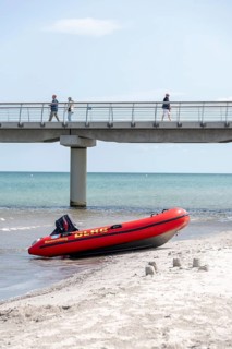 A DLRG lifeboat lies on the beach in front of a bridge with walkers and the sea in the background,
