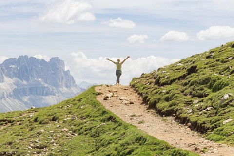 Person with outstretched arms on a path with a wide mountain view, Schlehrn hike, Alpe di Siusi,