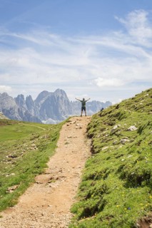 A hiker on a mountain path with outstretched arms in front of an impressive mountain scenery,