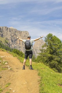 A man stands joyfully with outstretched arms on a sunny mountain path, Schlehrn hike, Alpe di