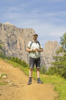 A man with a rucksack stands smiling on a mountain path with a rocky background, Schlehrn hike,