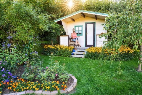A woman sits relaxed on the small veranda of a garden shed and reads. Blooming summer flowers in