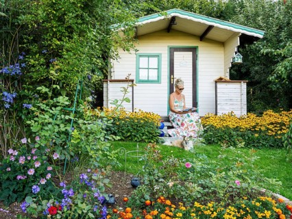 A woman in a dress sits relaxed on the small veranda of a garden shed and reads. Blooming summer