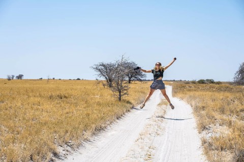 Tourist with camera jumping on a sandy track between yellow dry grass, Makgadikgadi salt pans,