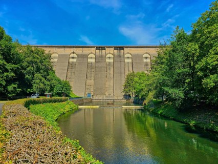 Large dam wall in the foreground, flanked by river and lush trees under a clear blue sky,