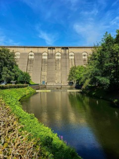 Dam wall with river in the foreground, surrounded by trees and reflected in the water,