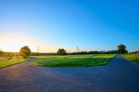 Fork of an asphalt road and a gravel path surrounded by a green meadow with wind turbines in the