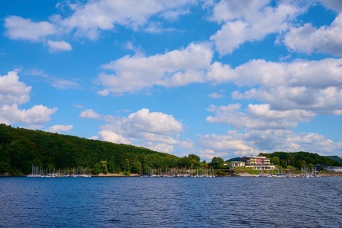 A lake with a building and boats along a wooded shore under a cloudy sky, summer, Rursee,