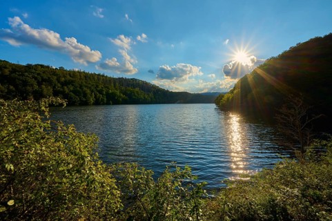 Sunbeams reflecting in the lake surrounded by wooded hills under a blue sky with clouds, summer,