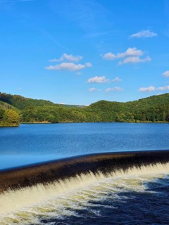 A quiet reservoir with a small waterfall, surrounded by wooded hills under a blue sky, summer,
