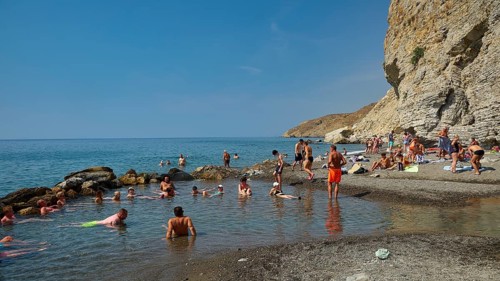 People relaxing on the beach in front of rocky coast and clear blue sea, Embros Thermal Baths, Kos,