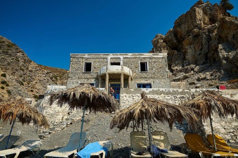 Stone house against a rocky backdrop with beach loungers and thatched roofs in the sunshine, Embros