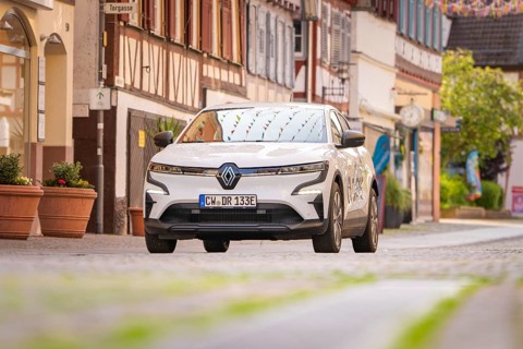 A white electric car drives through a narrow cobbled street with colourful shutters in the town,