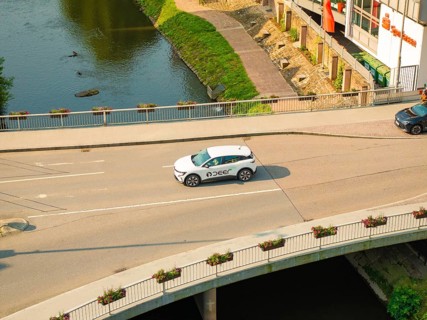 A white car drives on a bridge over a river, surrounded by urban architecture and nature, Deer E,