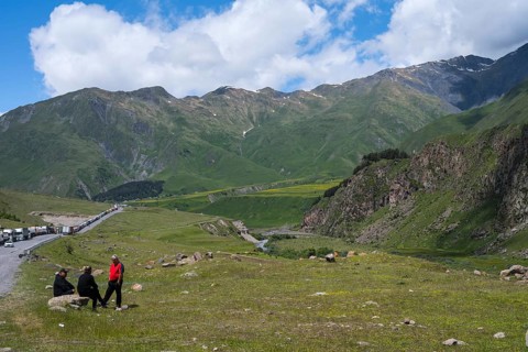 Three people sit in a meadow in front of a mountain range under a cloudy sky, left truck queue on