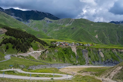 A small village is situated on a hill surrounded by green mountains and cloud-covered sky, views of
